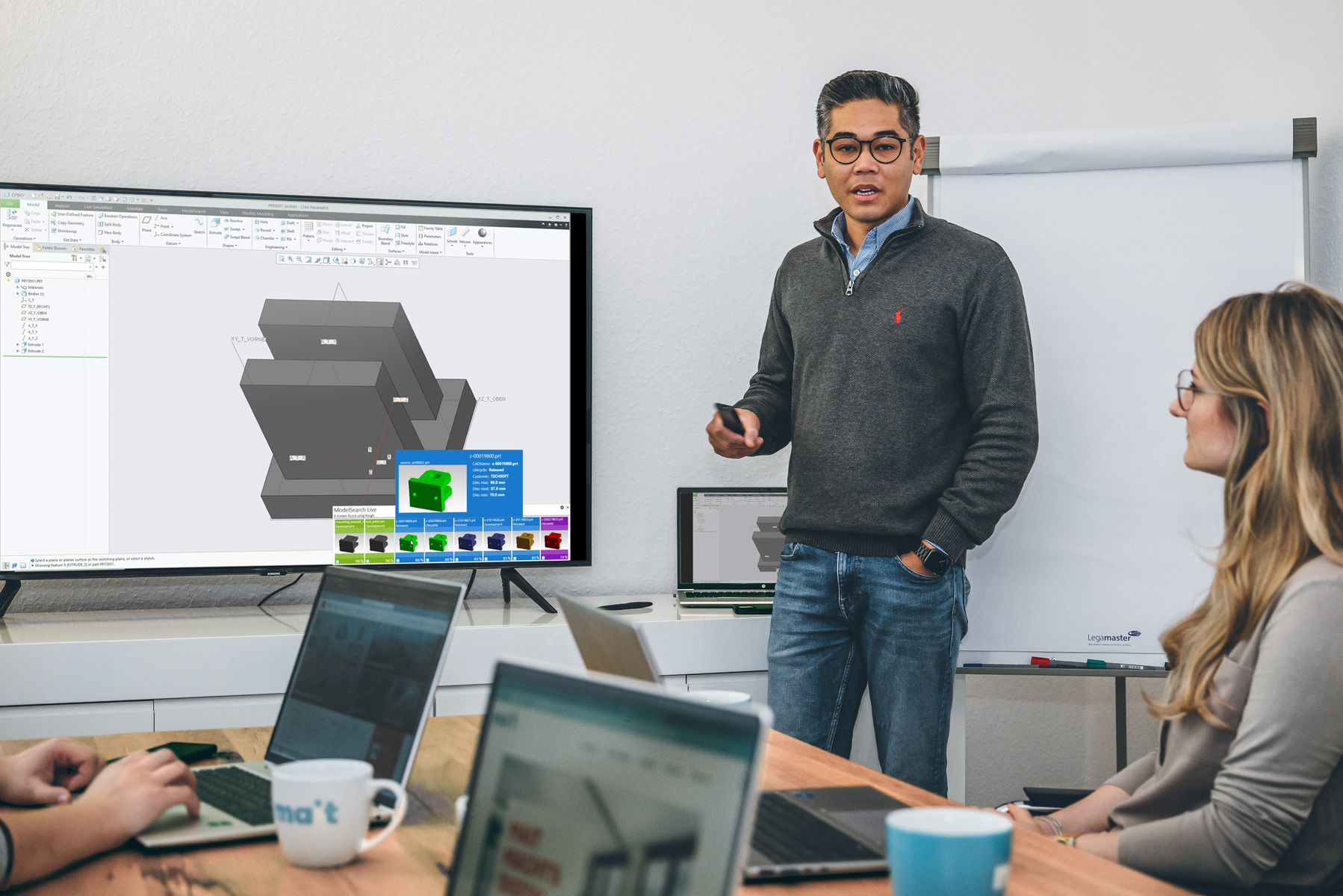 Person steht vor einem Bildschirm mit 3D-Modellierung und spricht zu einer Gruppe in einem Besprechungsraum mit Laptops und Flipchart.
